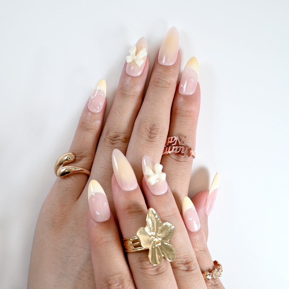 Handmade almond nails with soft ombre pink-yellow tips, pearls, and bow charm